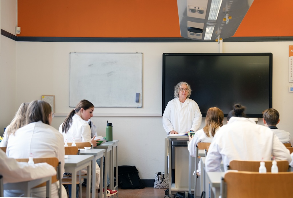 Docent staat voor het krijtbord in een klaslokaal gevuld met leerlingen in witte labjassen.