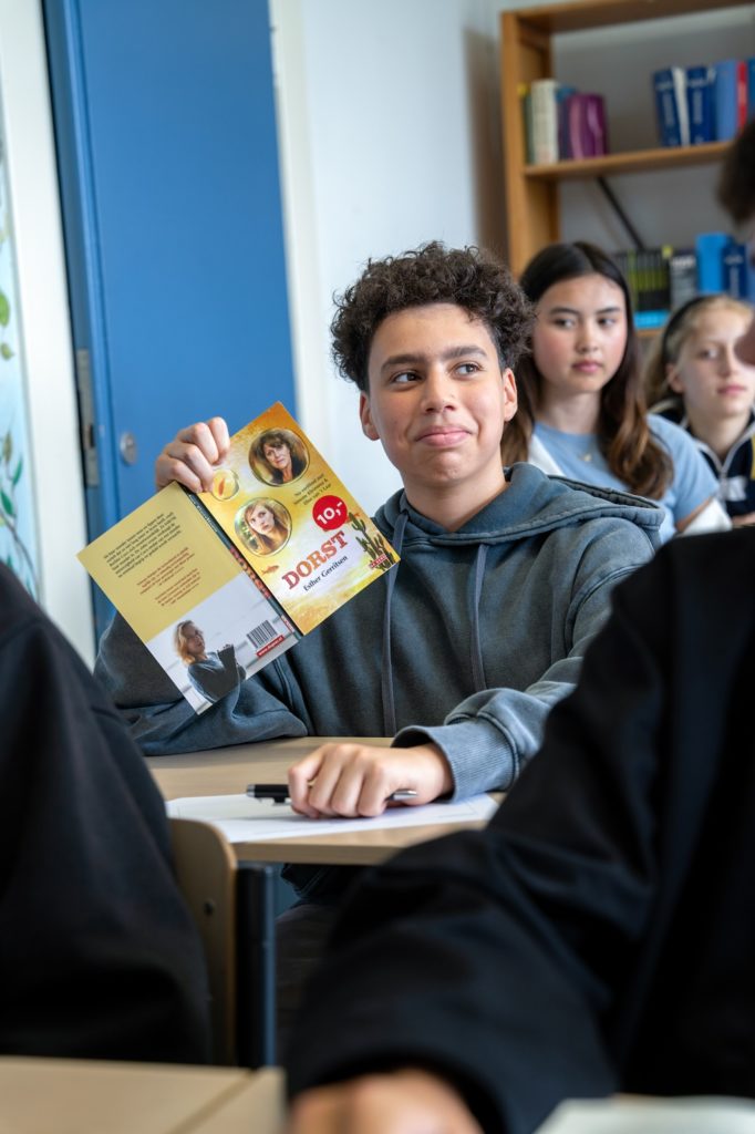 Jongen in de klas houdt zijn boek, dat is opengeslagen en een gele cover heeft, vast en kijkt een beetje sceptisch.