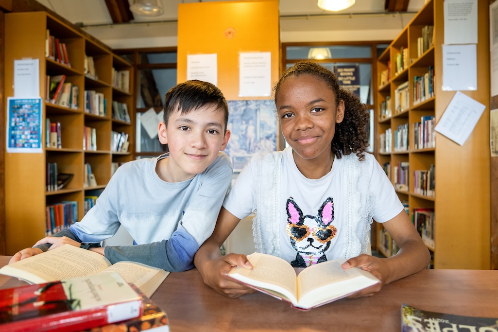 Twee kinderen die samen met een boek aan een tafel zitten en kijken in de camera.